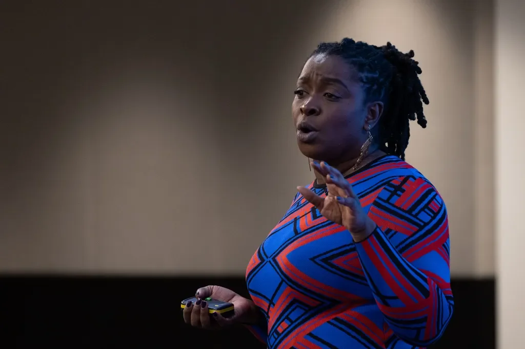 A woman with dark skin and dreadlocks wearing a colorful geometric patterned top, holding a remote control, speaking or gesturing with her hand.