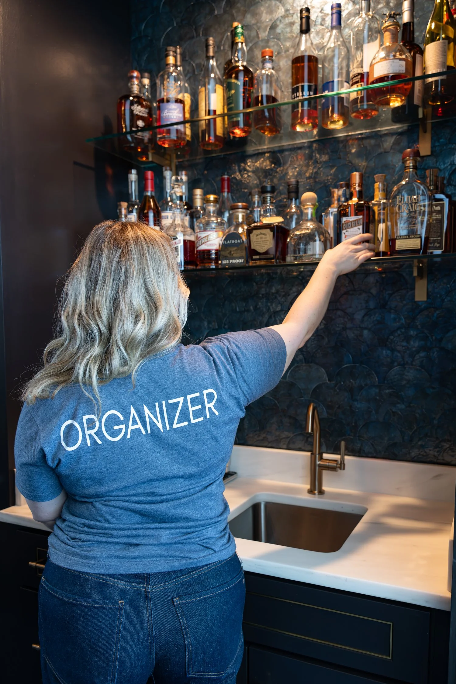 Woman with blonde hair stands in front of a glass bar full of rare bourbons.