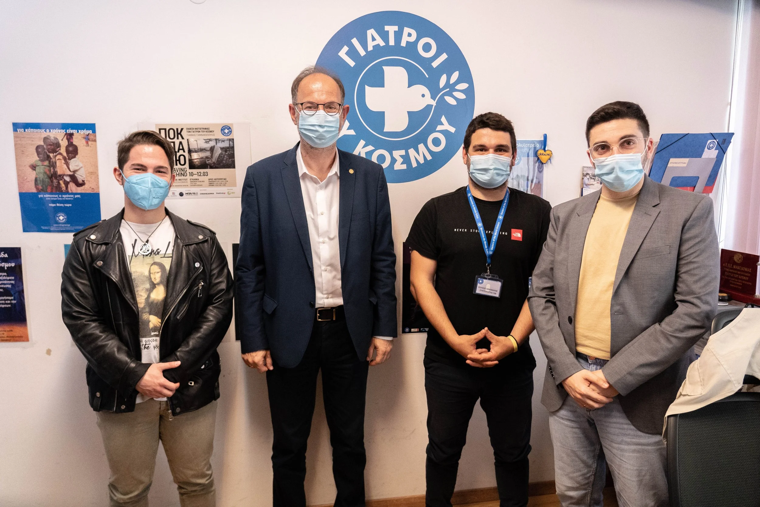 Four men wearing face masks standing in front of a wall with posters. A logo in Greek with a cross and dove is visible on the wall.