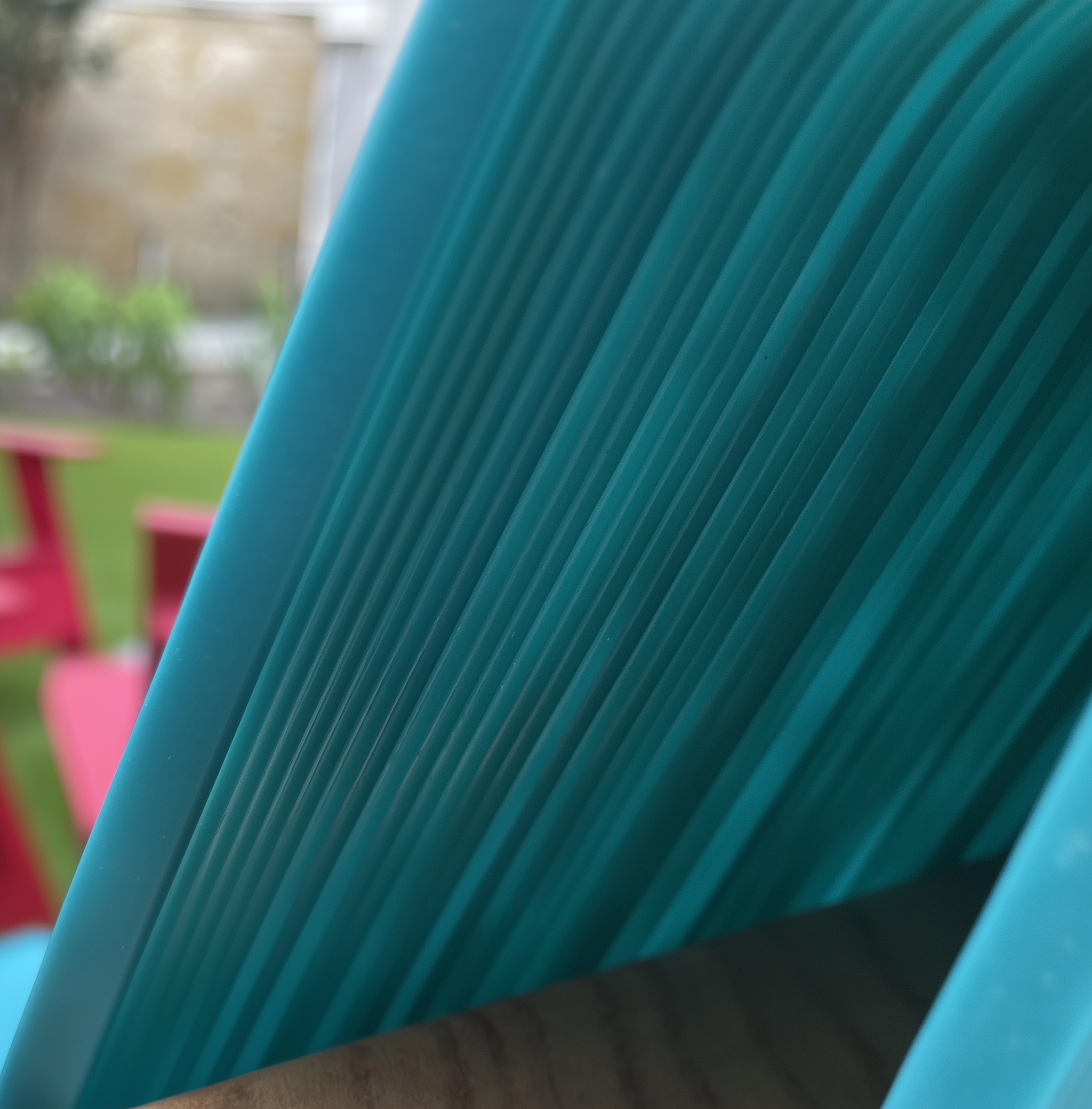 3D printed furniture - Close-up of a stack of blue plastic chairs outdoors, with blurred pink chairs and greenery in the background.