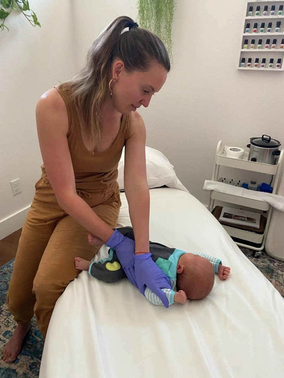 A woman wearing a brown sleeveless top and brown pants, with purple gloves, is gently holding a baby on a white examination table in a medical setting.
