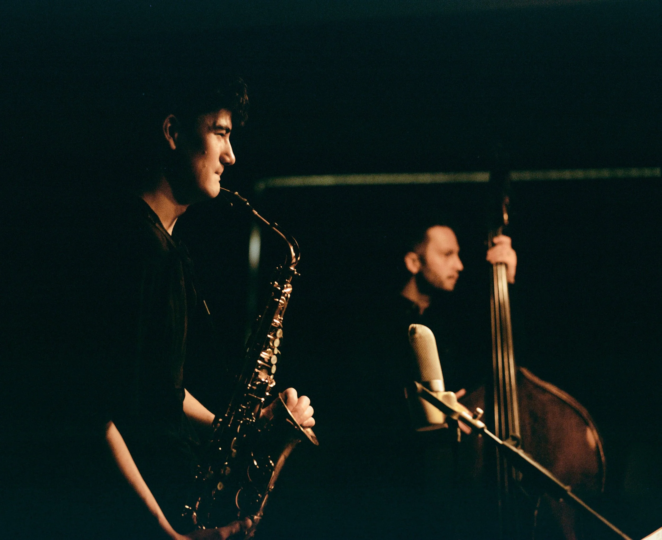 Zwei Musiker spielen in einem dunklen Raum, einer mit Saxophon, der andere mit Kontrabass, vor einem Mikrofon.