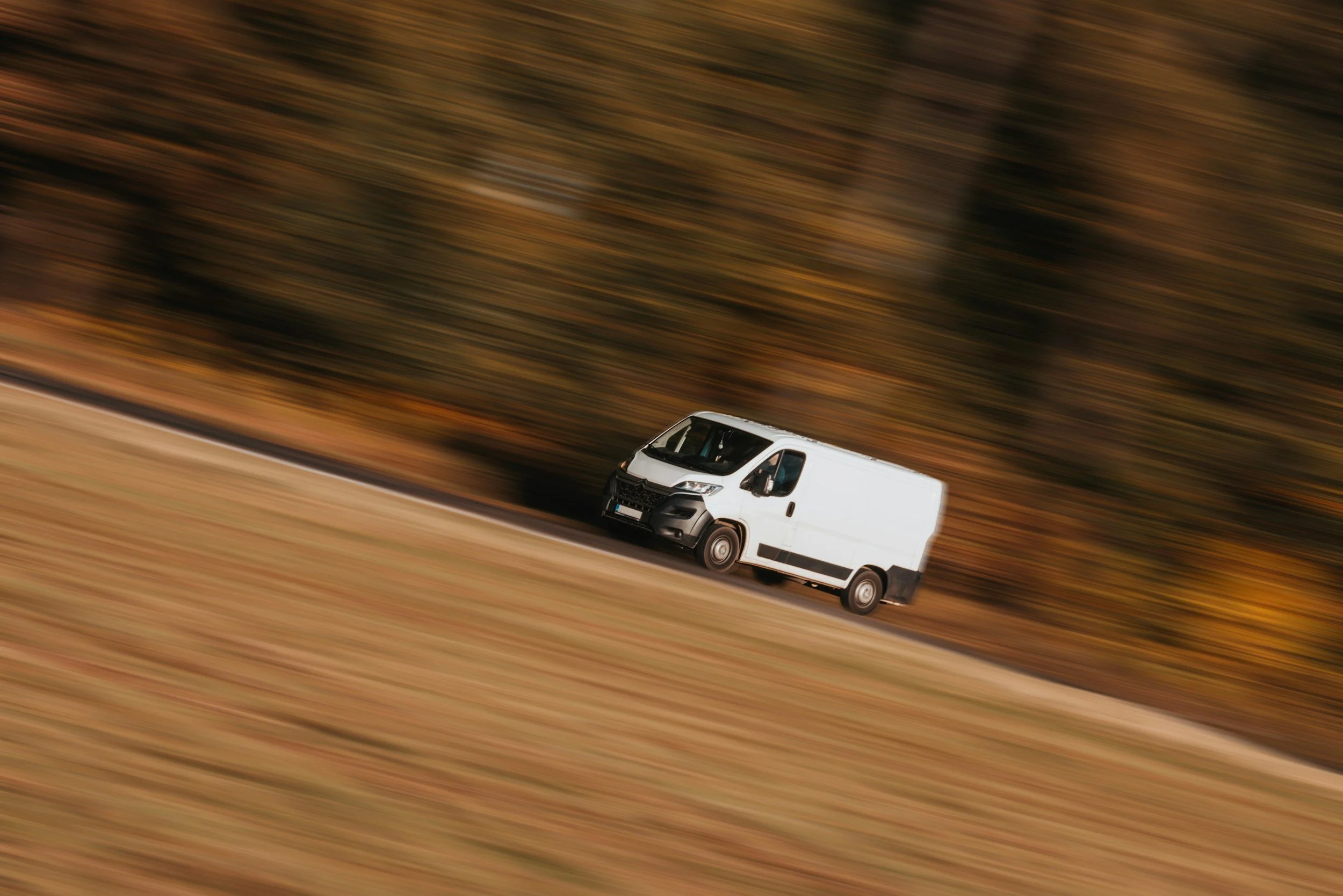 A white delivery van moving quickly on a highway during autumn, with blurred brown and orange trees in the background.