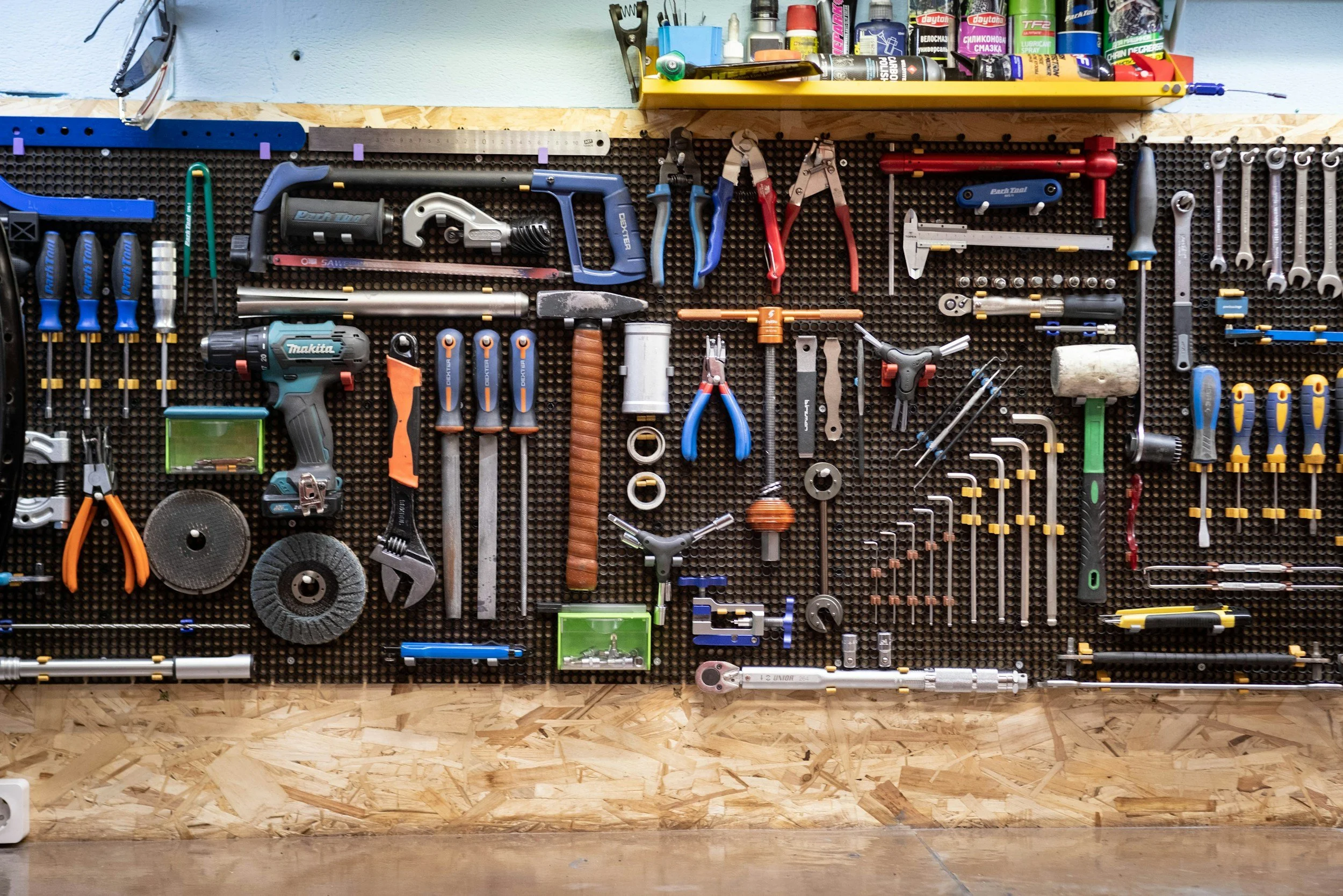 Pegboard with various hand tools such as screwdrivers, pliers, wrenches, a cordless drill, a hammer, and measuring tools, organized on a workshop wall. A yellow shelf with cans, bottles, and various maintenance supplies is above the pegboard.
