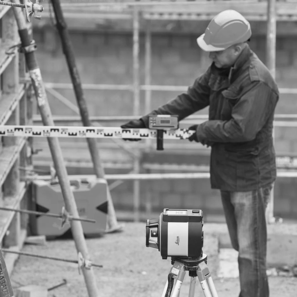 Construction worker wearing a helmet and jacket using a surveying instrument on a construction site.