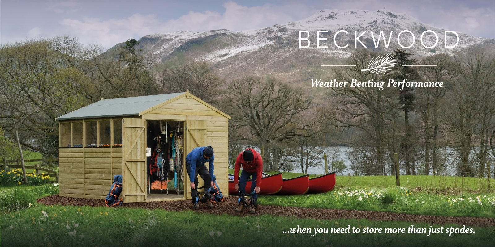 Two people are standing outside a wooden shed, packing backpacks. The shed is located on a grassy area with trees and a lake in the background, with snow-capped mountains behind. Two red canoes are on the ground near the shed. The sky is cloudy.