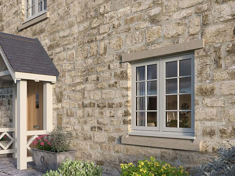 Close-up of a stone house exterior with a gray-framed window, a small porch with a gabled roof, and potted plants and flowers in front.