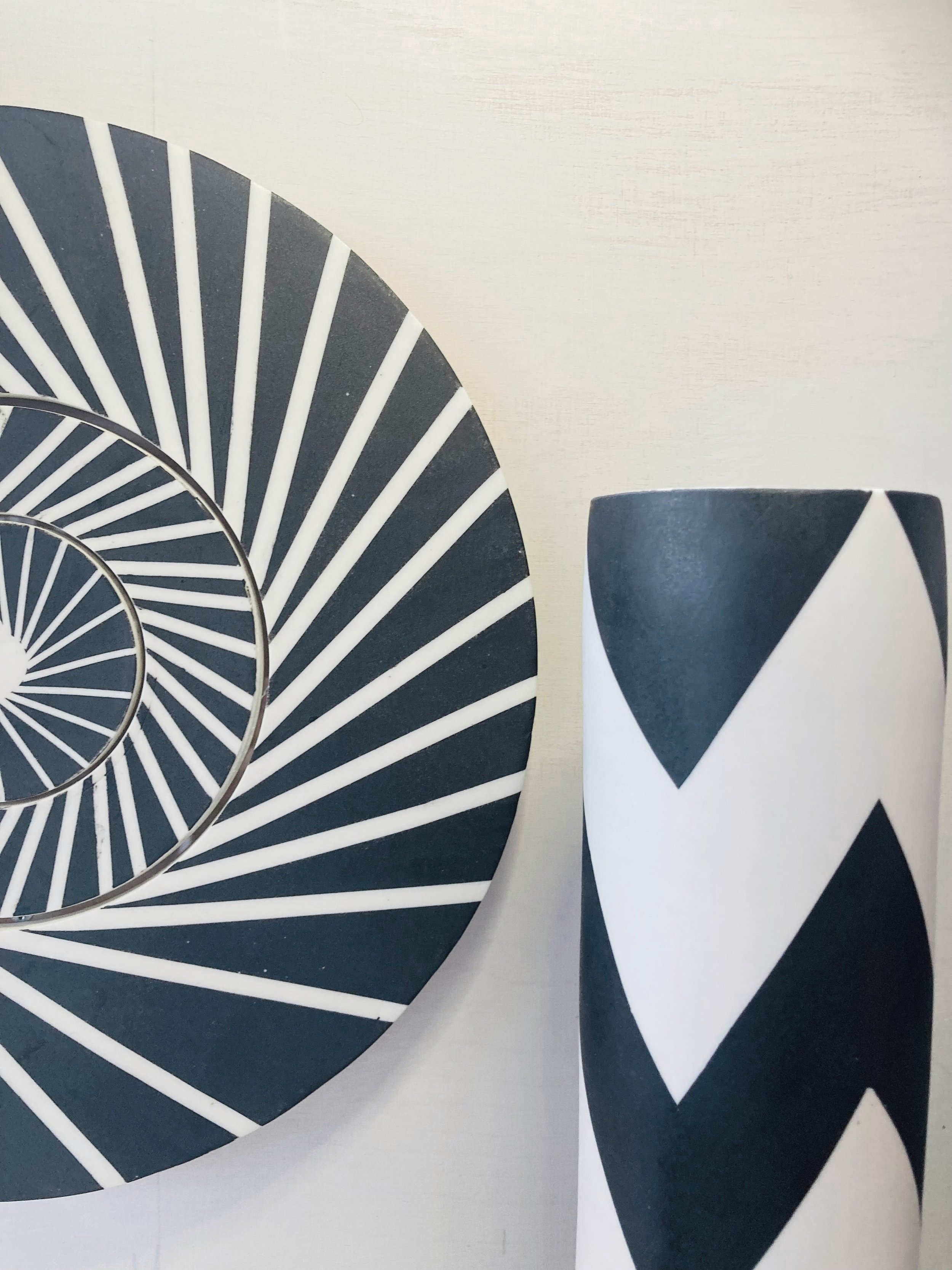Disc shaped wall piece made of concentric circles decorated with radiating black and white stripes to create a piece that creates the illusion of movement. This is presented alongside a zigzag cylinder vase