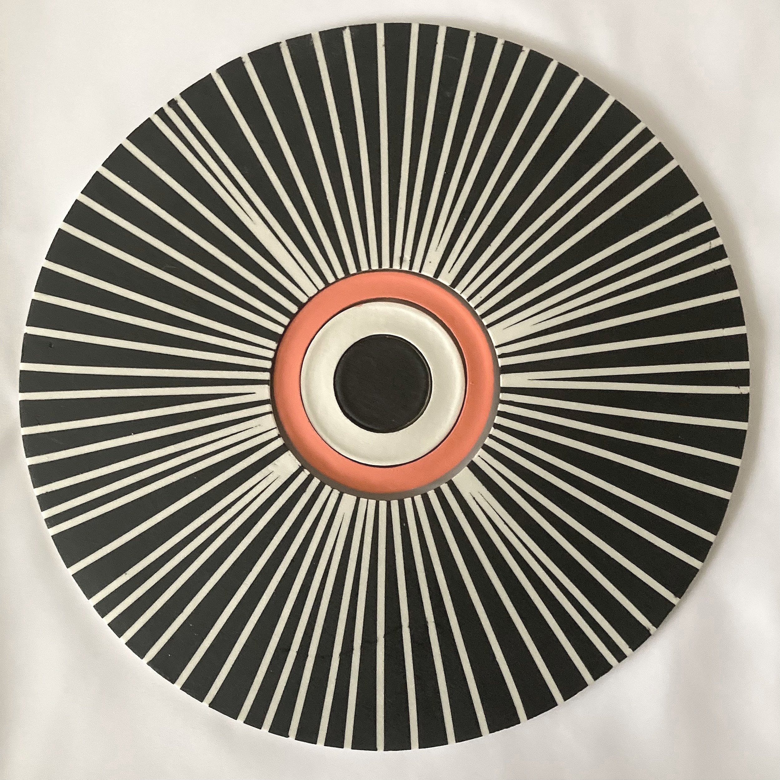 Disc shaped wall piece in porcelain made of concentric circles showing black white and orange centre with radiating black and white lines on the outside