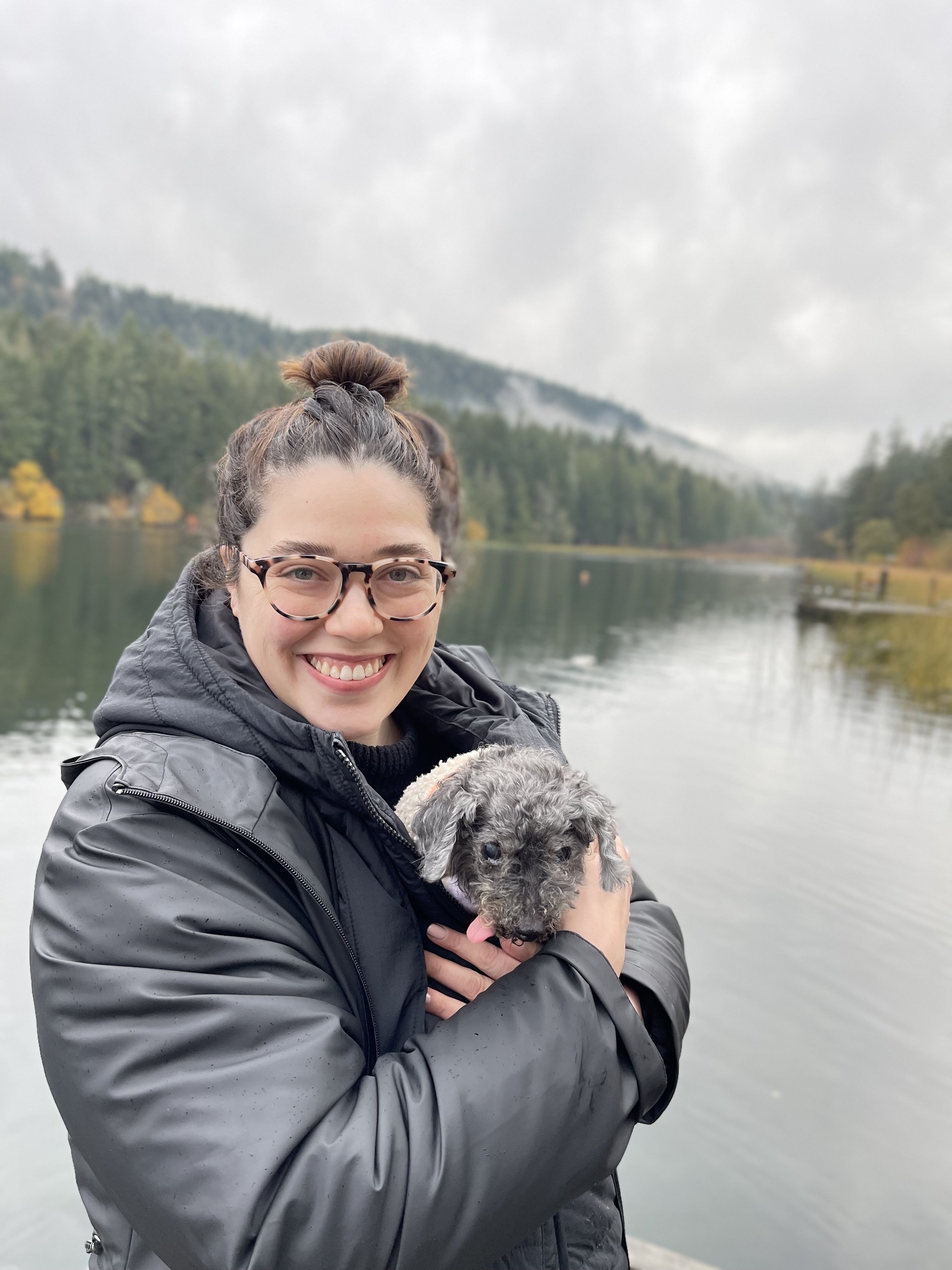A woman in a black jacket and glasses smiling while holding a small gray puppy with pink tongue in front of a lake and forested mountains under cloudy sky.