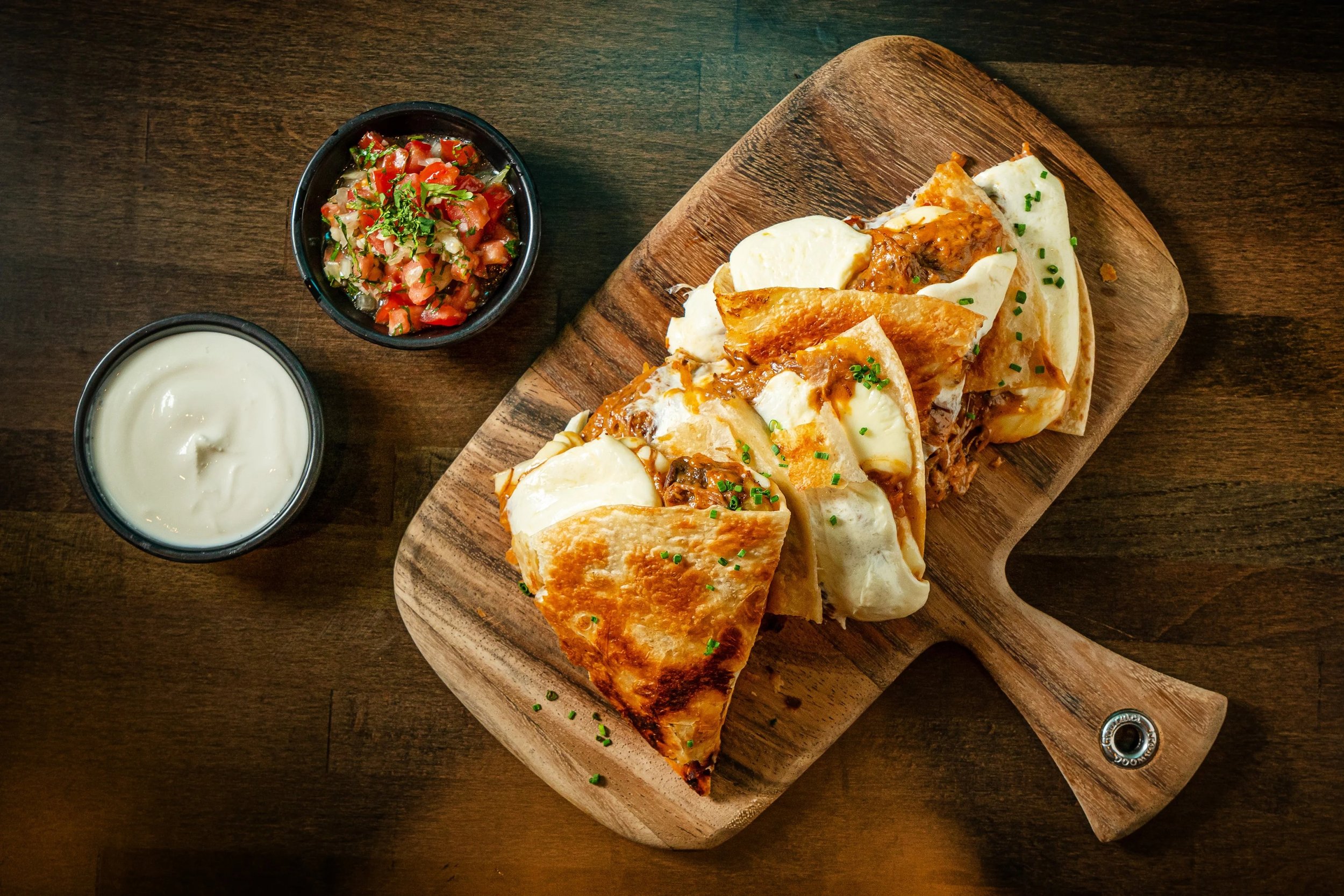 A wooden serving board with three tacos topped with chopped chives and cheese, accompanied by a small bowl of salsa and a small bowl of sour cream on a dark wooden table.