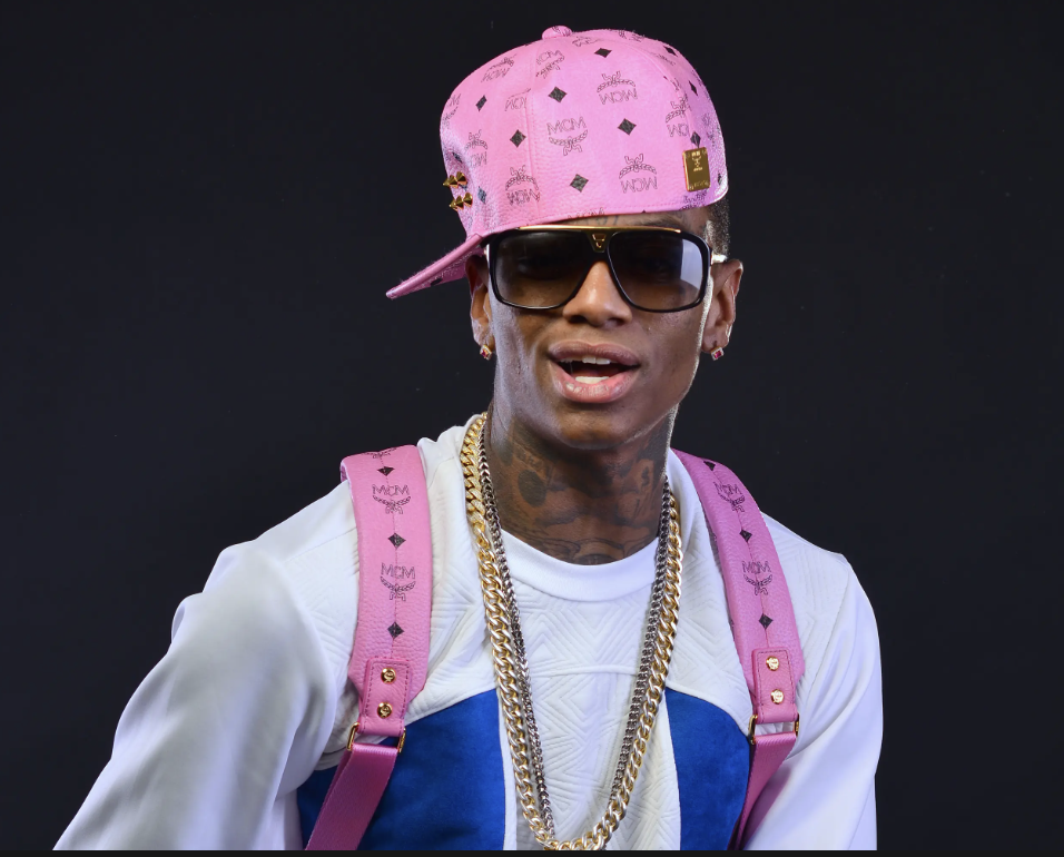 A man wearing a pink cap, sunglasses, white and blue athletic jacket, pink backpack with gold accents, and gold chains.