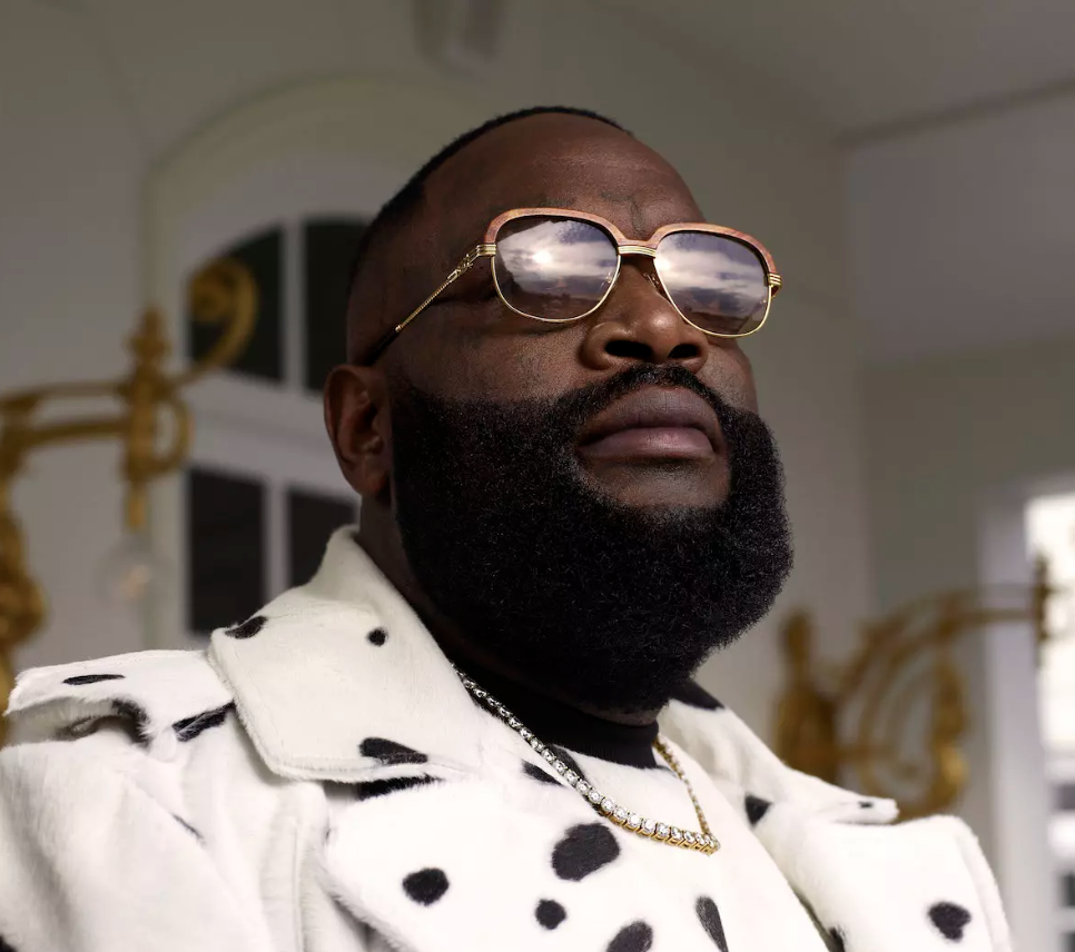 A man with a full beard and glasses, wearing a white jacket with black spots, inside a room with ornate gold decorations.