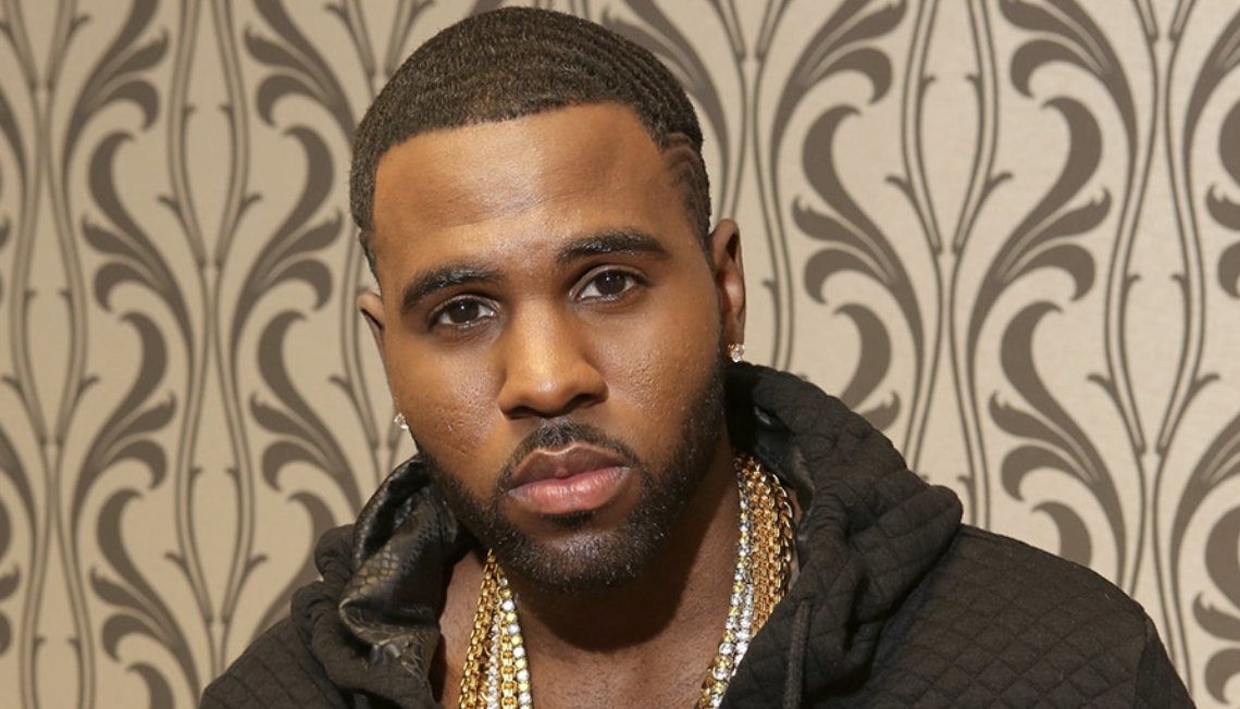 Close-up of a man with short hair, diamond earrings, multiple gold chains, wearing a black hoodie, standing in front of a patterned wallpaper.