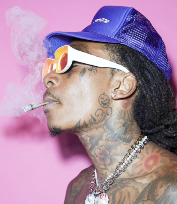 A tattooed man wearing a blue cap, white sunglasses with orange lenses, and jewelry, smoking a joint with pink background.