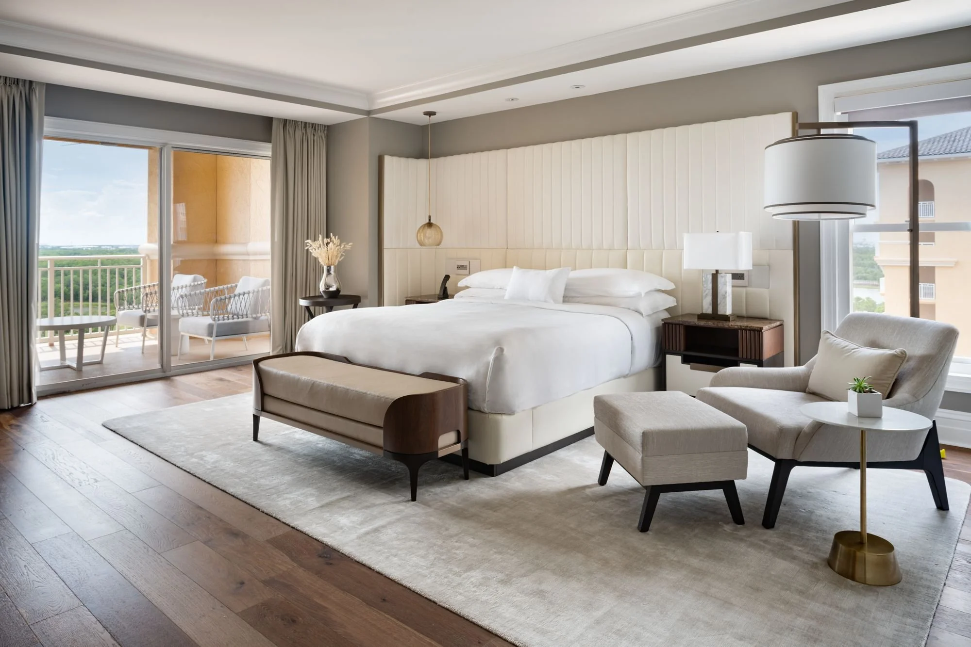 A modern hotel room with a large bed, sitting area, and balcony with outdoor seating and a view of greenery.