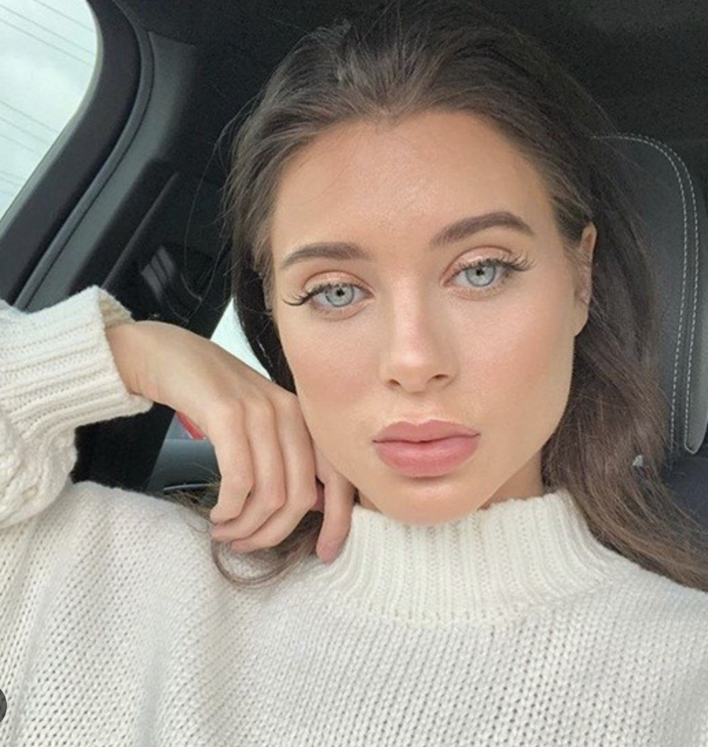 A woman with blue eyes and brunette hair taking a selfie inside a car, resting her head on her hand, wearing a white knit sweater.