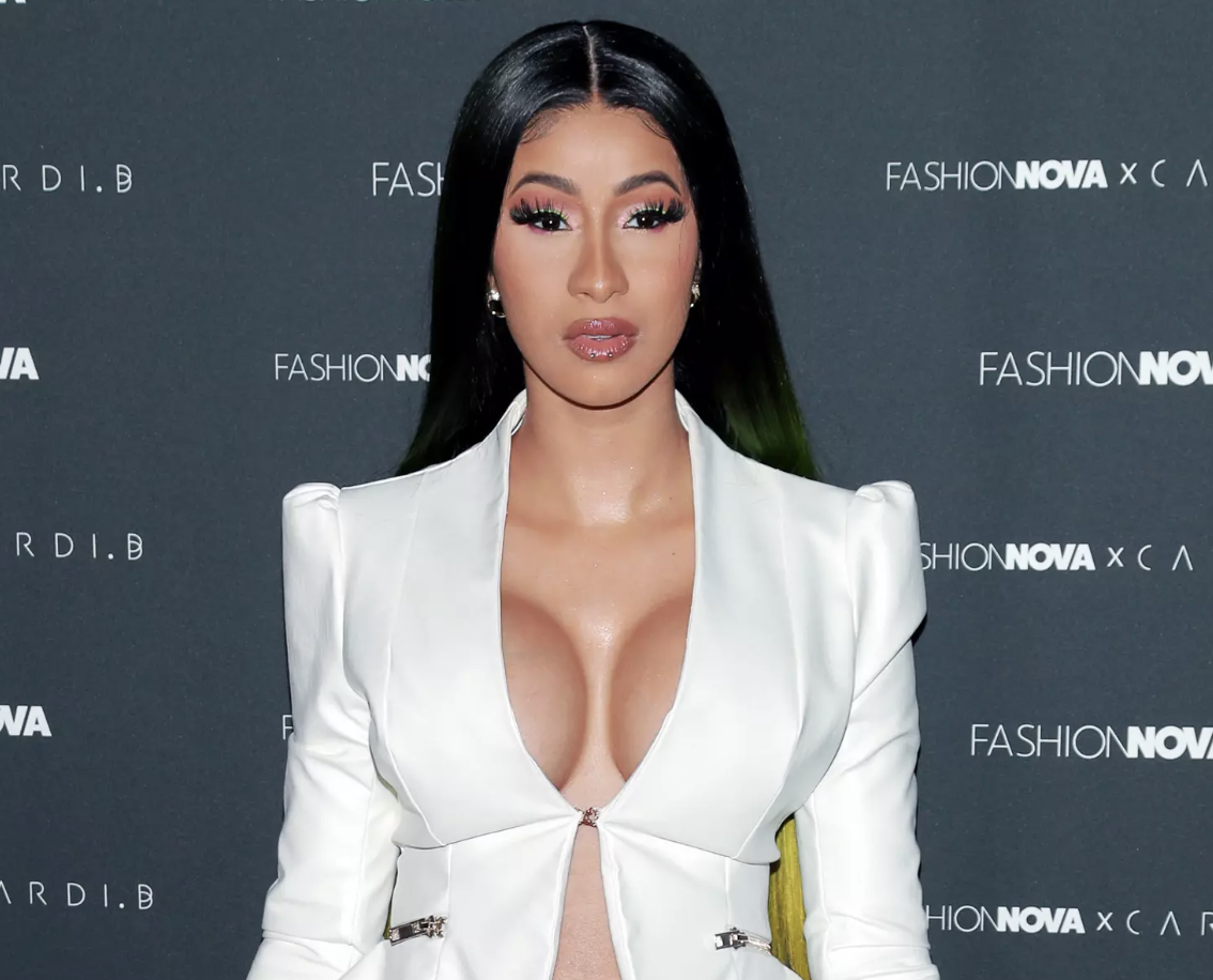 A woman with long black and green hair posing in front of a dark backdrop with the words "Fashion Nova" printed on it, wearing a white blazer with a plunging neckline.
