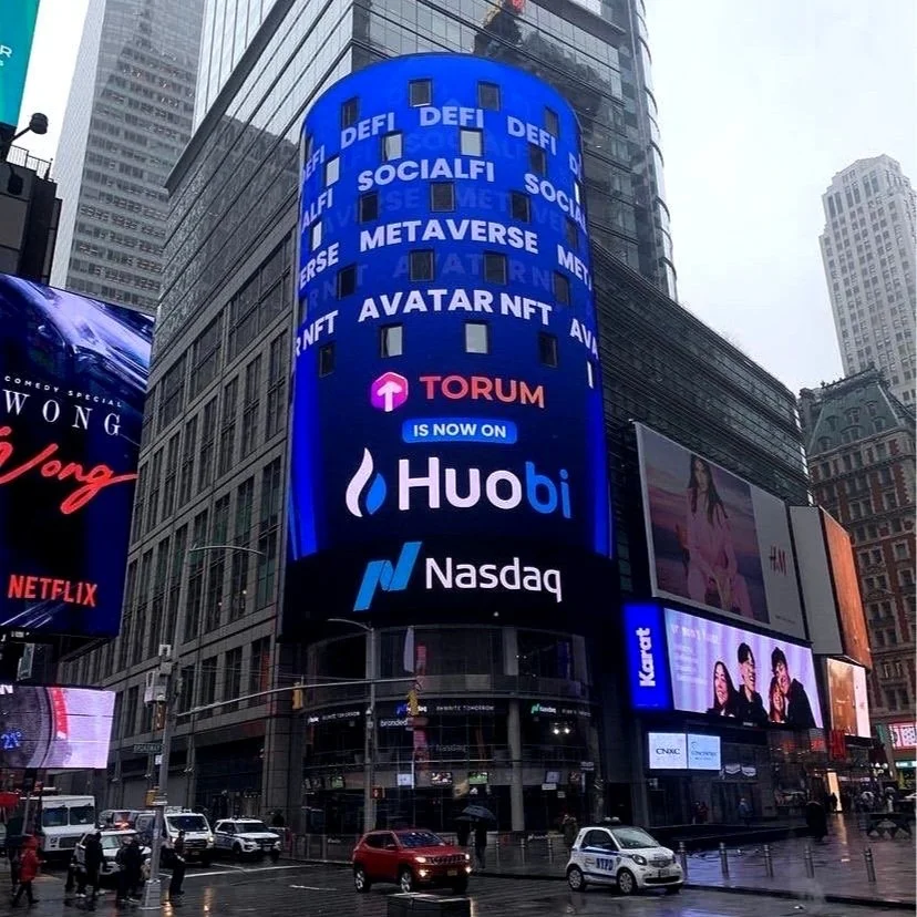 Digital billboard in Times Square featuring advertisements for various cryptocurrencies and platforms, including Huobi, Nasdaq, and Torum, with people and vehicles on the street below on a rainy day.