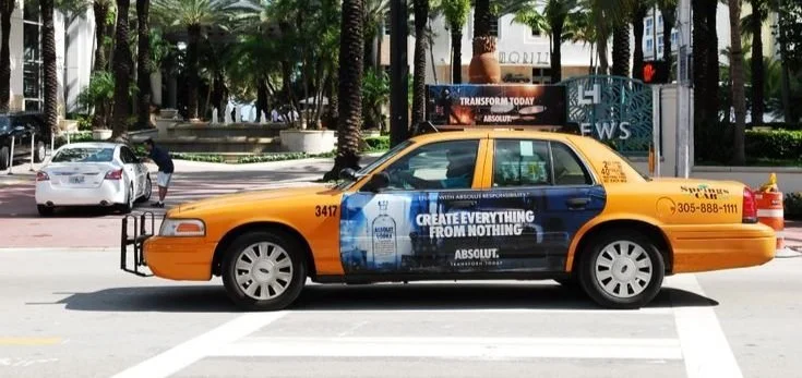 Yellow taxi cab with an advertisement on the side, parked on a city street with palm trees in the background.