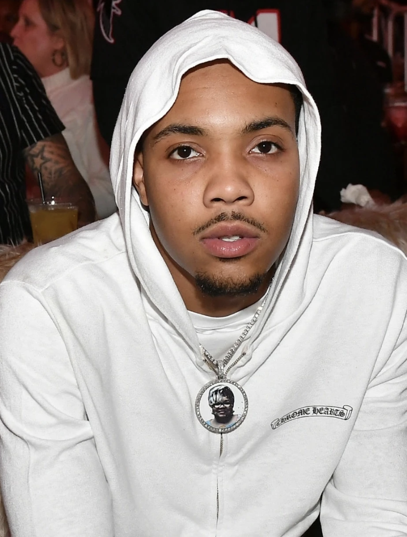 Young man with a hoodie and a chain necklace featuring a pendant with a young person's face, sitting indoors with a blurred background of other people and drinks.