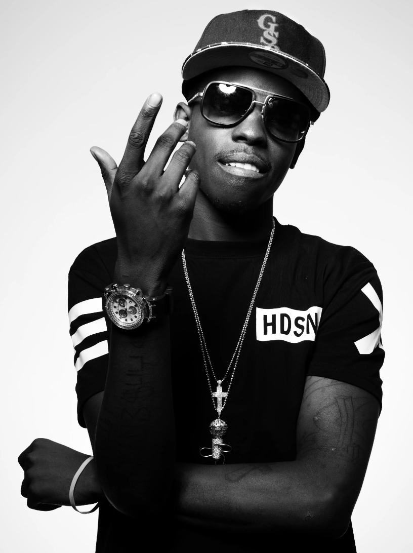 Black and white photo of a young man wearing sunglasses, a baseball cap, a black T-shirt with white stripes on sleeves and 'HDSN' printed on chest, multiple necklaces, a watch, and a bracelet. He is making a gesture with his right hand and has tattoos on his left arm.
