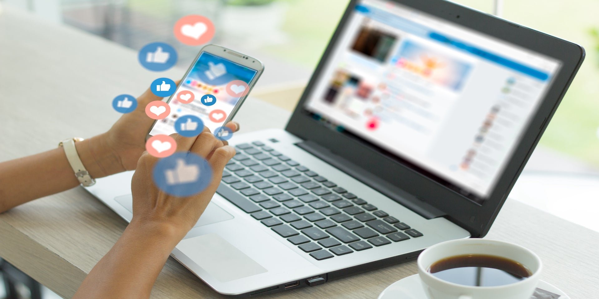 Person using smartphone with floating social media icons in front of laptop on a desk with coffee cup.
