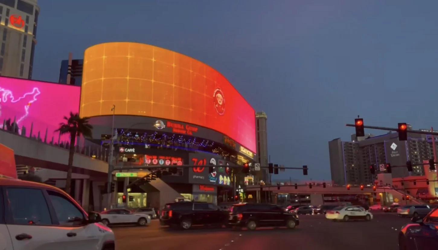 Digital billboards and neon signs in a busy city intersection at dusk, with cars stopped at red lights and tall buildings in the background.