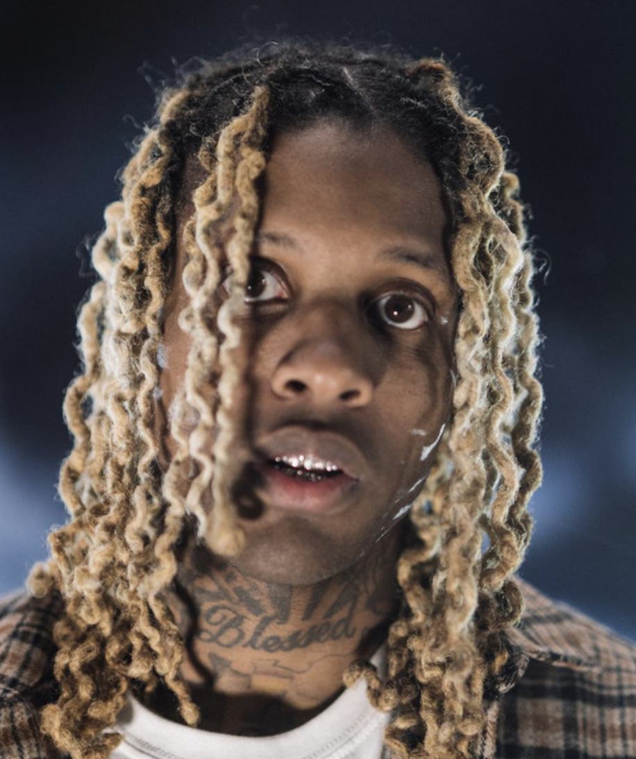 Close-up of a young man with dreadlocks, tattoos, and a surprised expression, standing in a dark environment.