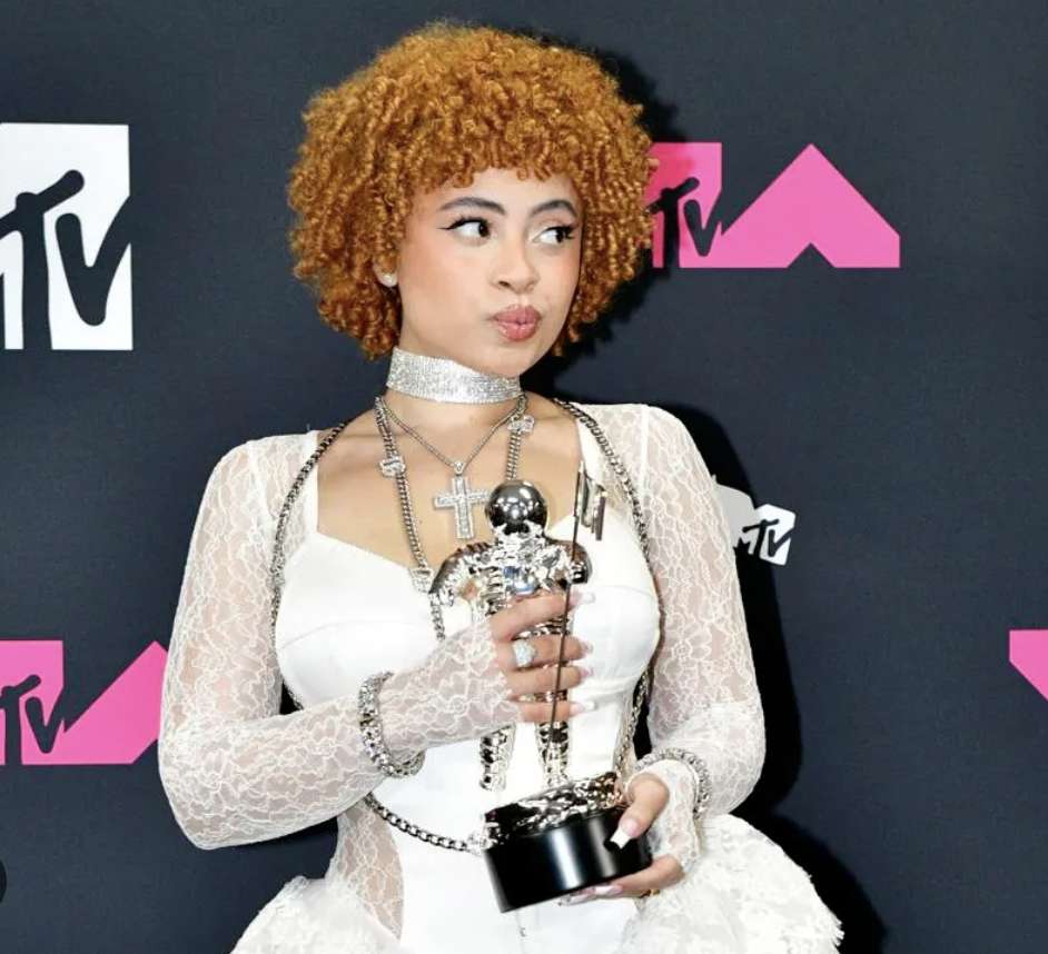 Young woman with curly orange hair in a white lace dress and silver jewelry holding an MTV award statue.