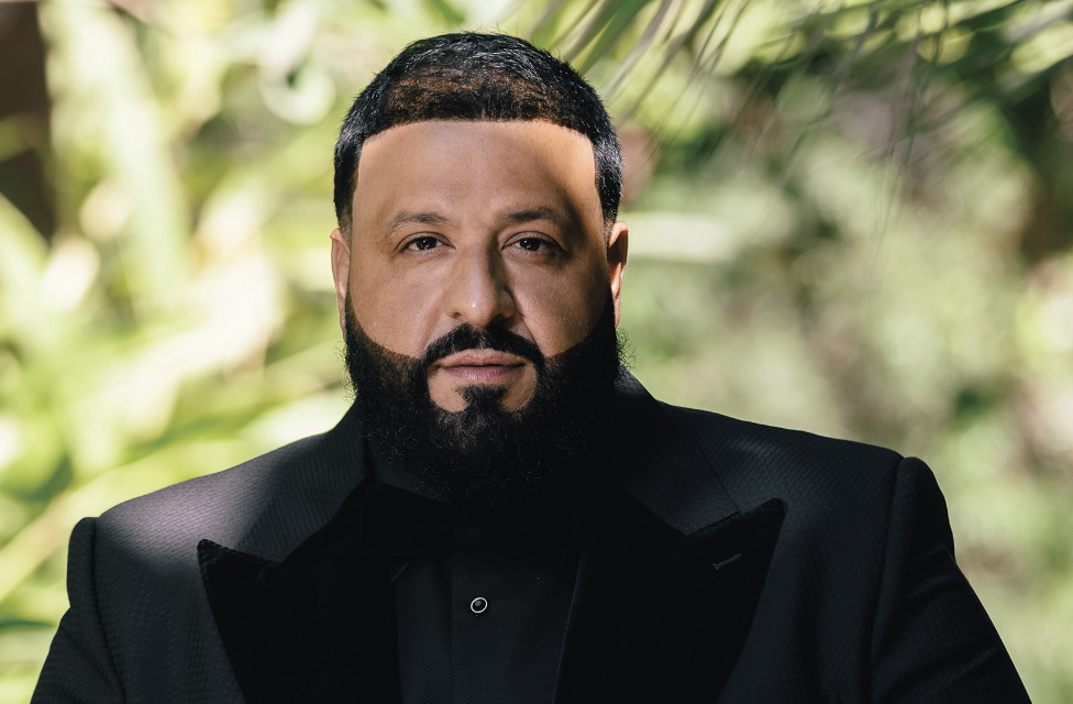 A man with a well-groomed beard and short hair, wearing a black tuxedo, standing outdoors with greenery in the background.