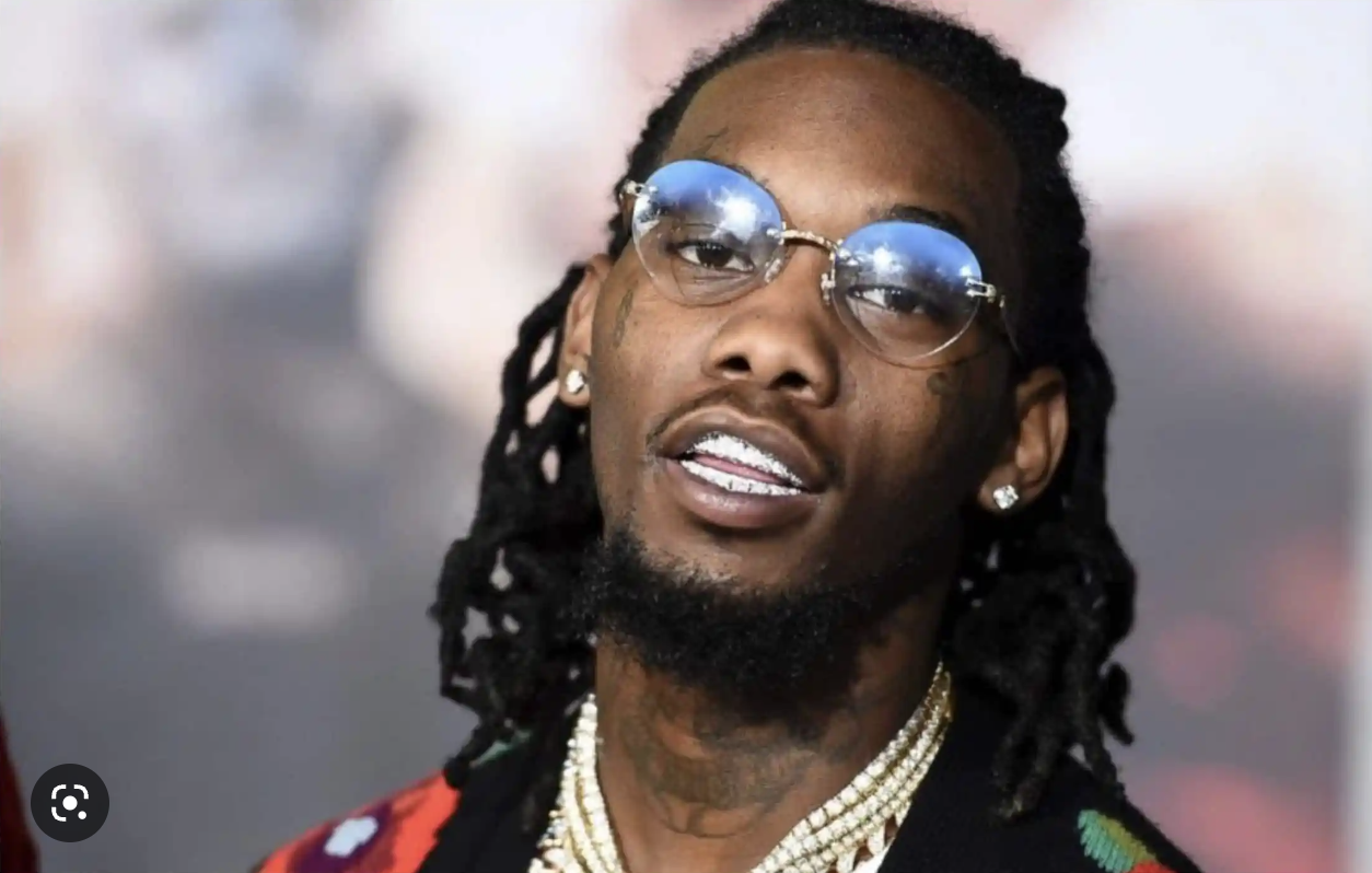A man with long dreadlocks wearing round sunglasses, diamond earrings, a chain necklace, and a grill showing his teeth, with a blurred background.