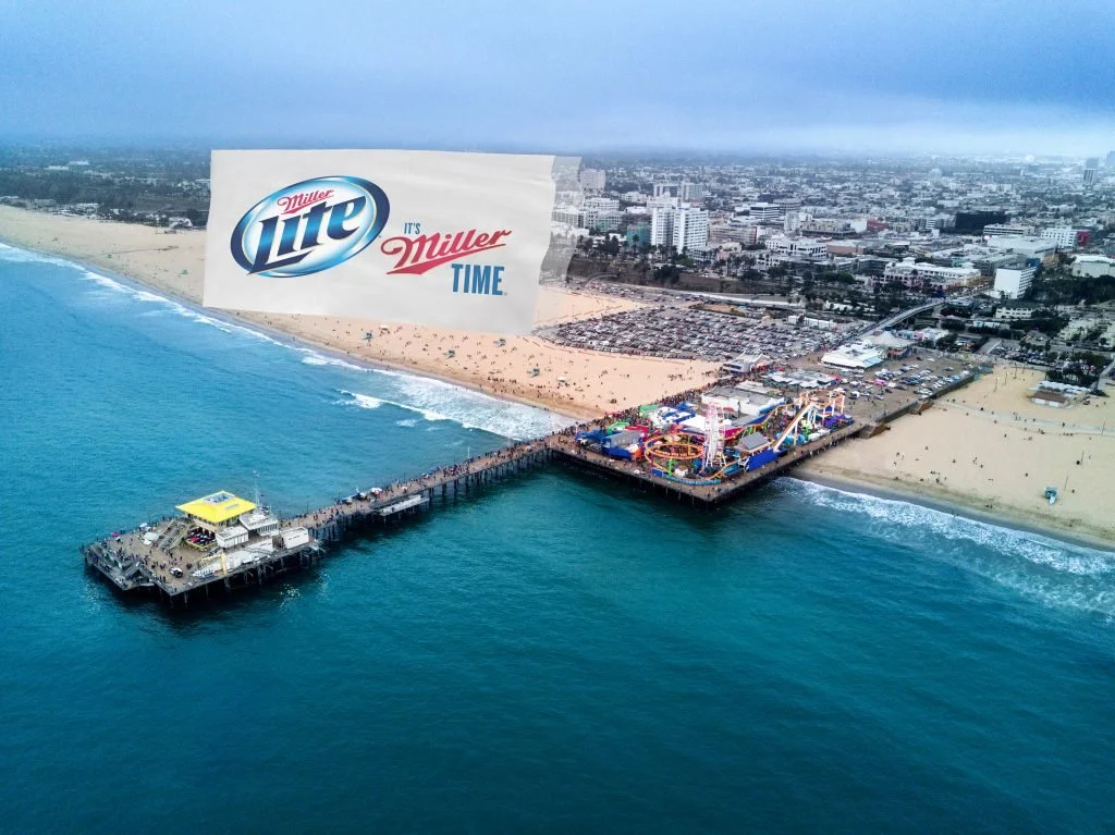 Aerial view of a beachside amusement park with a pier extending into the ocean, featuring rides and tents, and a cityscape in the background. A large Miller Lite advertisement floats over the scene.