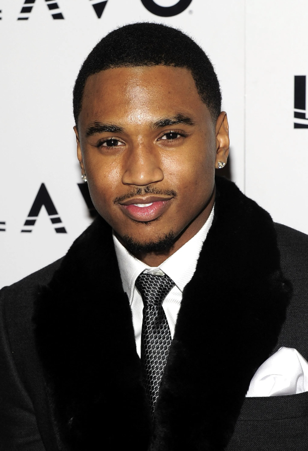A young man with short black hair, wearing a black suit with a white shirt, a patterned black and white tie, and a white pocket square. He has earrings and is smiling slightly at the camera. The background features white with black text and graphics.