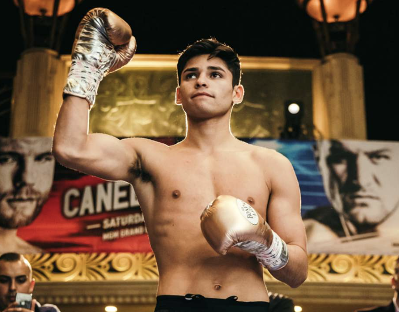 A young male boxer with dark hair, shirtless and wearing gold boxing gloves, posing with his right arm raised in a fighting stance at a weigh-in event. In the background, there are promotional posters for a boxing match.