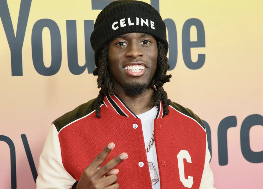 Young man smiling, wearing a black beanie with 'CELINE' written on it, a red and white varsity jacket, and a white shirt, making a peace sign with his right hand, standing in front of a colorful background with the YouTube logo.