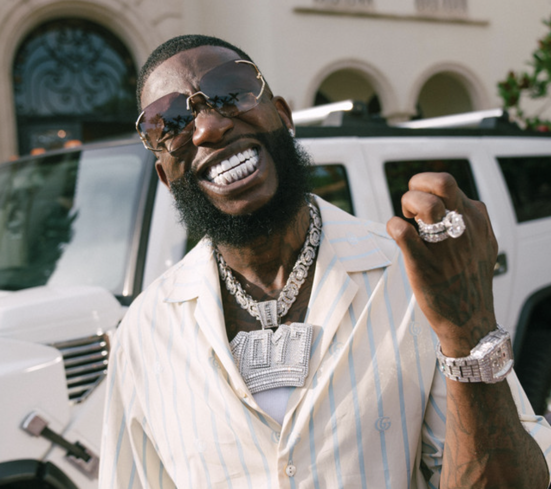 A man with a beard and sunglasses, smiling and showing his teeth, wearing a light-colored, striped shirt, jewelry including rings, necklaces, and a watch, and clenched his fist in front of a white vehicle, in a sunny outdoor setting.