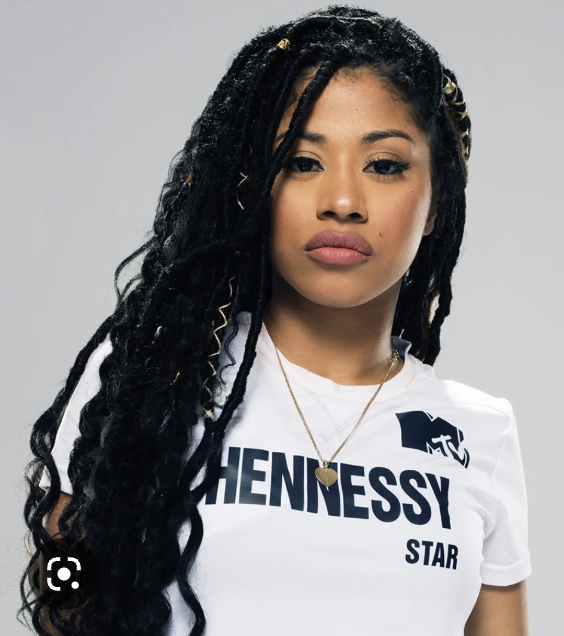 Young woman with long, curly black hair and a serious expression wearing a white t-shirt with Hennessy logo and gold necklace, against a plain background.