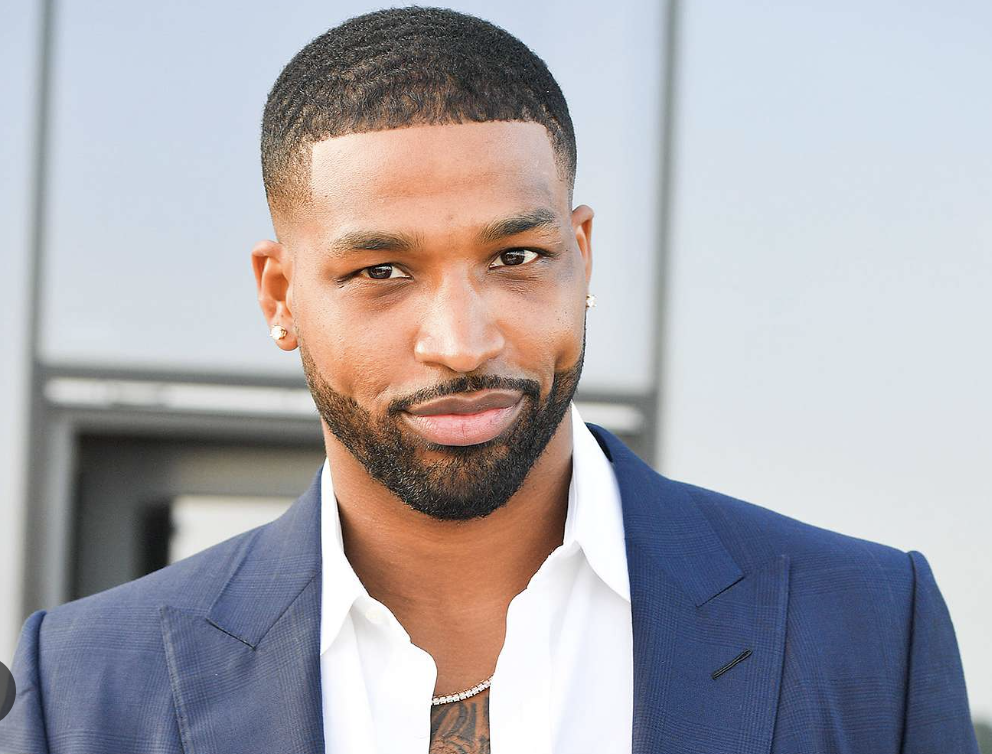 A confident young man with a trimmed beard and short hairstyle, wearing a navy blazer over a white shirt, standing in front of a building with a gray background.