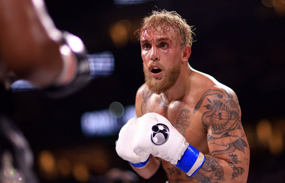 A shirtless male boxer with tattoos on his chest and arms, and with blood on his face, is in a boxing ring, wearing white gloves with black logos, preparing to fight an opponent whose arm is visible holding a water bottle.