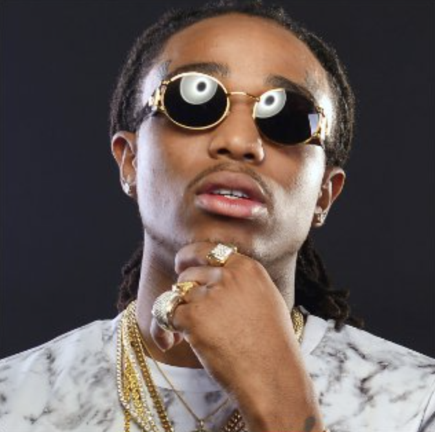 A person wearing gold-rimmed sunglasses, multiple gold chains, rings, and earrings, with a thoughtful pose against a dark background.
