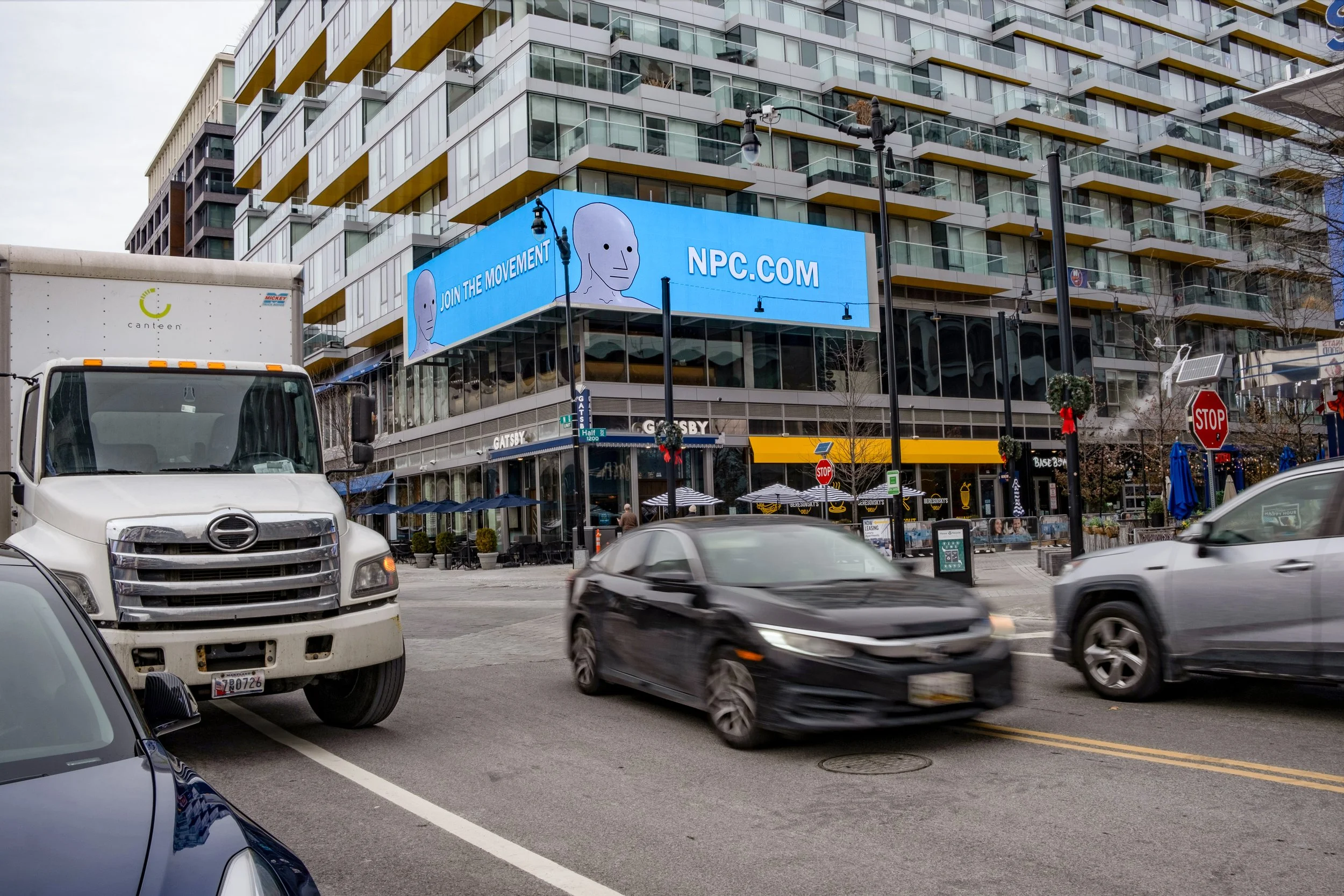 A city street with cars driving and parked, a large building with a digital billboard displaying an NPC character and the text ‘JOIN THE MOVEMENT NPC.COM’, and outdoor seating at a café underneath the billboard.