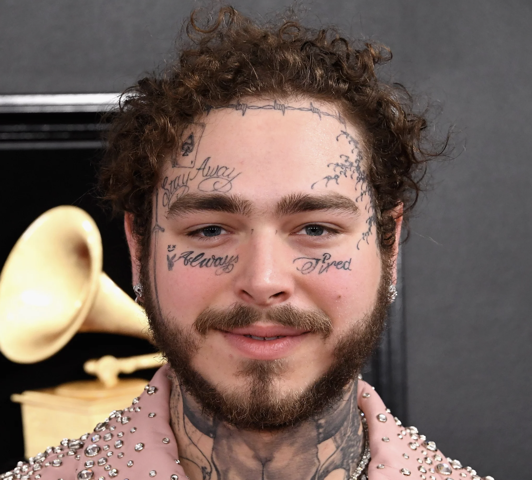 A young man with curly hair and facial tattoos smiling in front of a black background with a gold gramophone.