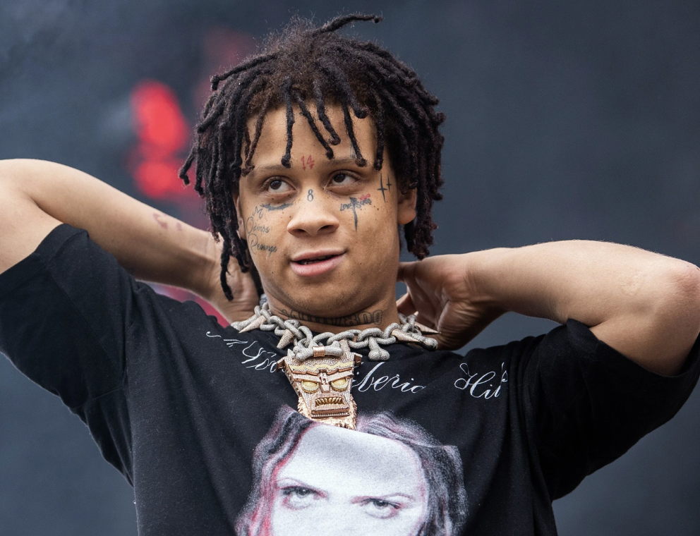 A young man with dreadlocks and tattoos on his face, wearing a black T-shirt with a graphic print, a chunky chain necklace, and a large metallic pendant, standing with his hands behind his head.