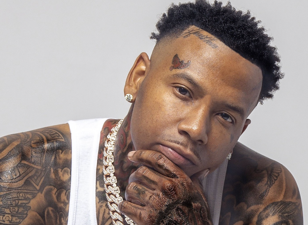 Close-up of a tattooed man with short curly hair, wearing a white sleeveless shirt, diamond earrings, and a chunky diamond chain, posing with his hand on his chin against a plain background.