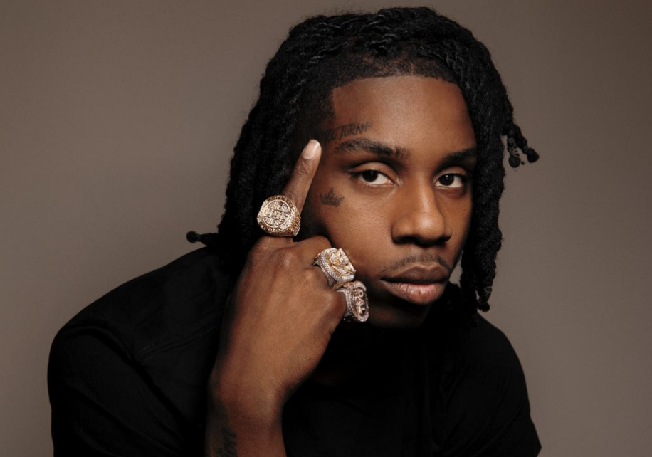 Close-up portrait of a young man with dreadlocks, wearing multiple large rings, resting his finger on his face, against a neutral background.