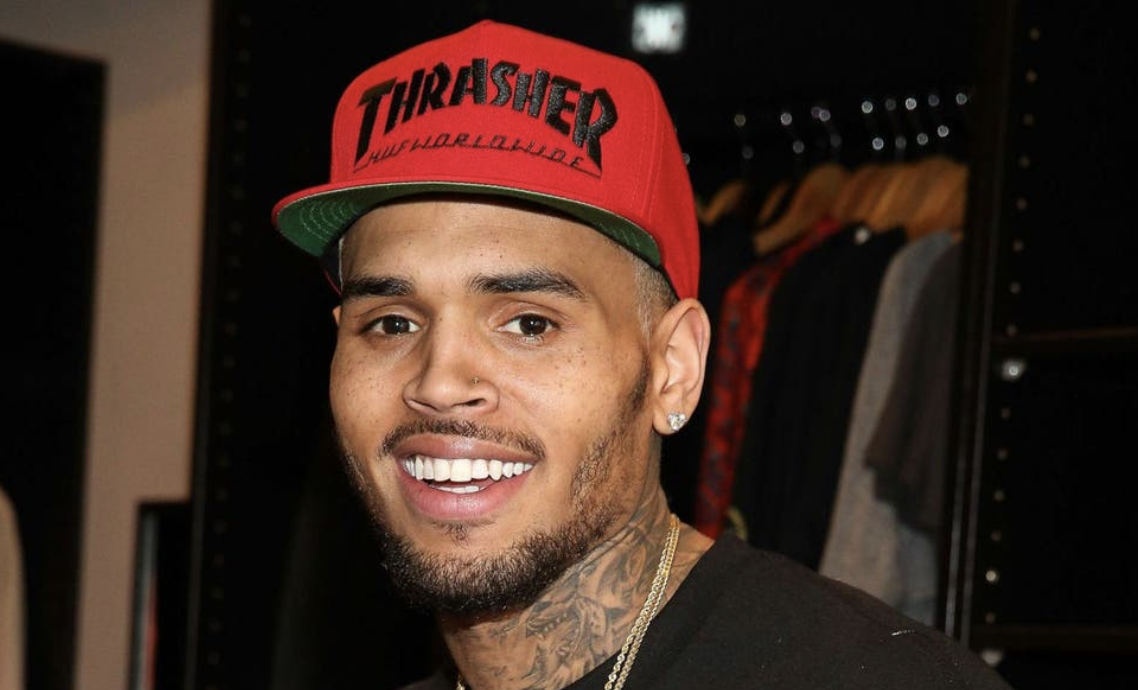 Close-up of a smiling man wearing a red Thrasher baseball cap, earrings, and a necklace, with tattoos visible on his neck, in front of a clothing display.