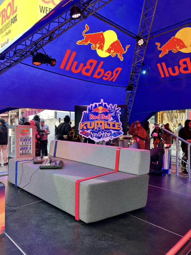 A stage set up for the Red Bull Kumite event in New York, under a large blue tent with the Red Bull logo. There is a gray sofa with red accents in the foreground, a digital screen displaying the event logo, and a crowd of people in the background.
