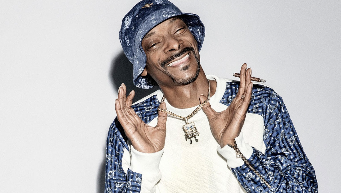 Man wearing a blue bucket hat, white sweater, patterned blue and white jacket, with jewelry, smiling, holding a cigar in one hand, posing against a plain background.