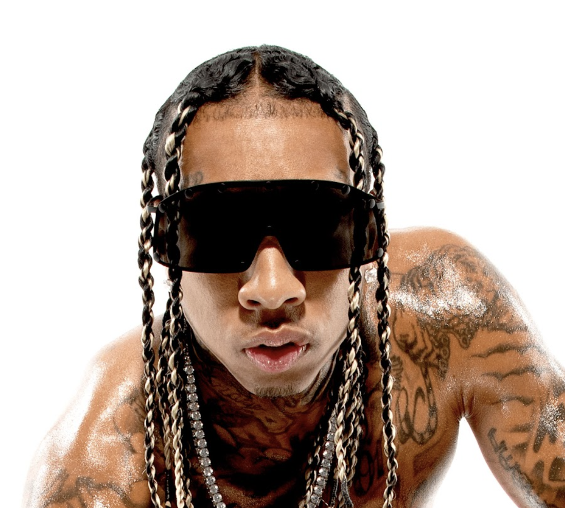 Close-up of a shirtless man with tattoos, wearing large black sunglasses and with long braids, against a white background.